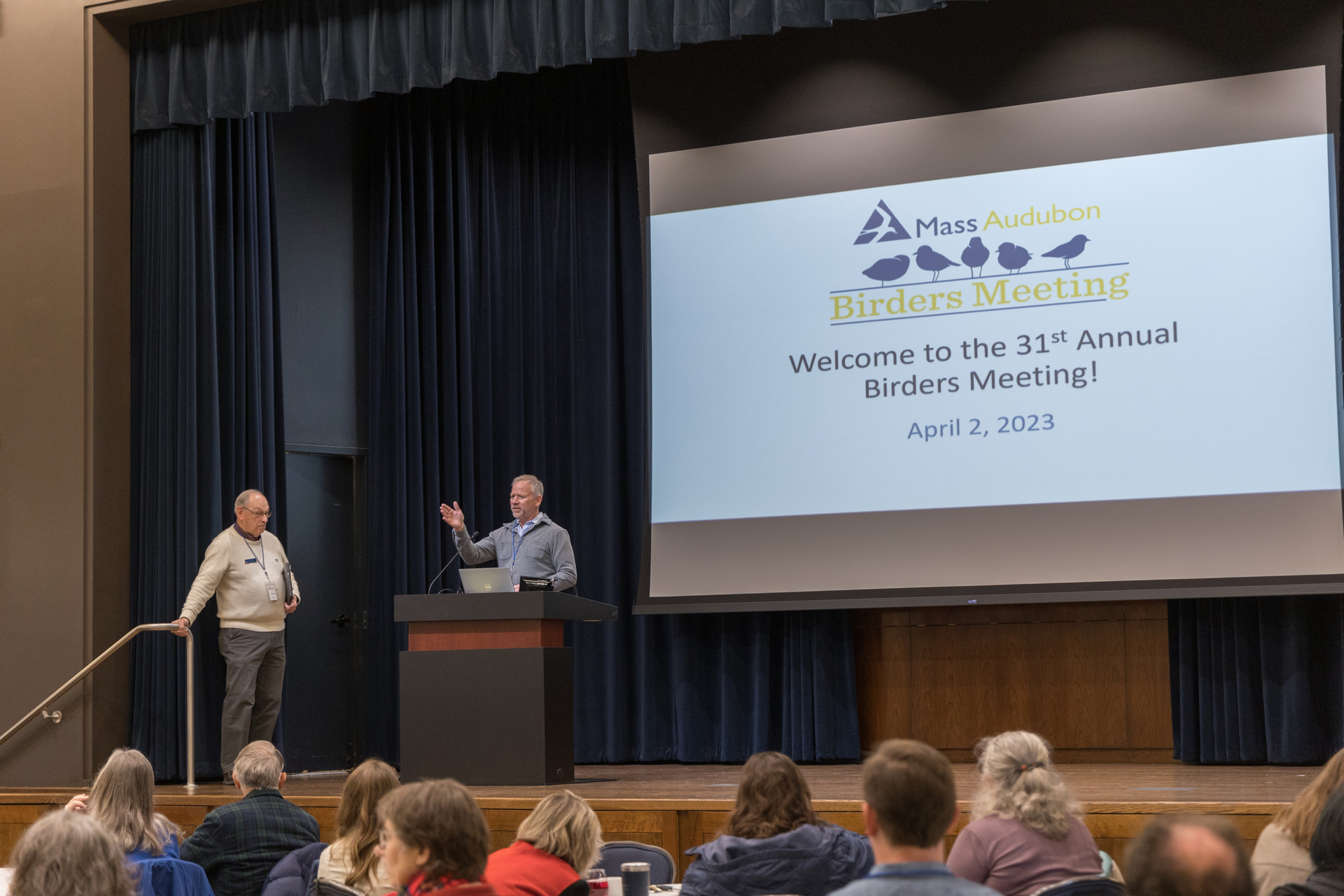 David O'Neill and Wayne Petersen on a stage in front of audience. on big screen slide that says 2023 Birders Meeting