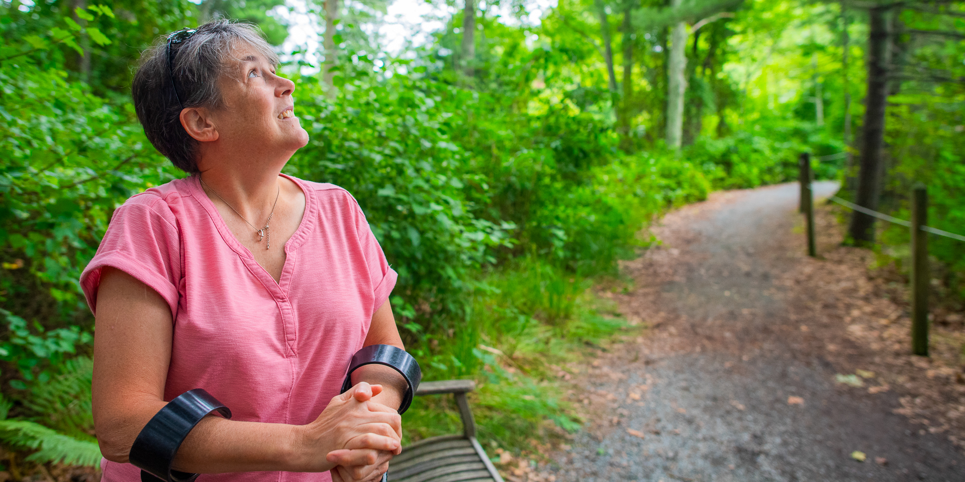 A woman wearing a pink shirt and using forearm crutches pauses to rest and look for birds in the forest canopy at a bench along Stony Brook's wheelchair-accessible All Persons Trail