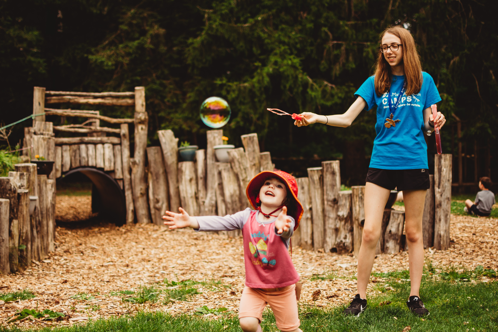 A counselor at Moose Hill Nature Camp makes bubbles while a camper chases after them in the Nature Play Area