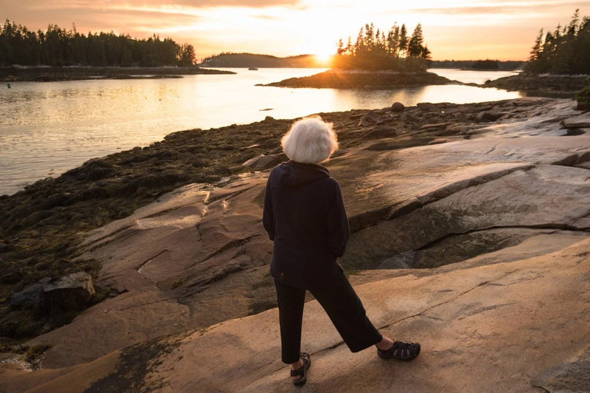 Person with gray-white hair standing on rocks looking at the setting sun © Joshua Touster