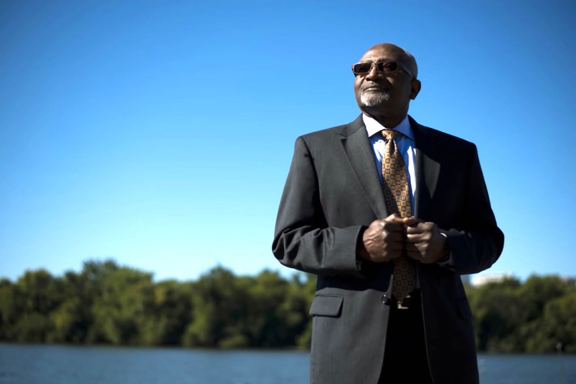 A portrait of Robert Bullard wearing a suit and sunglasses, standing in front of a pond edged with green trees under a clear blue sky