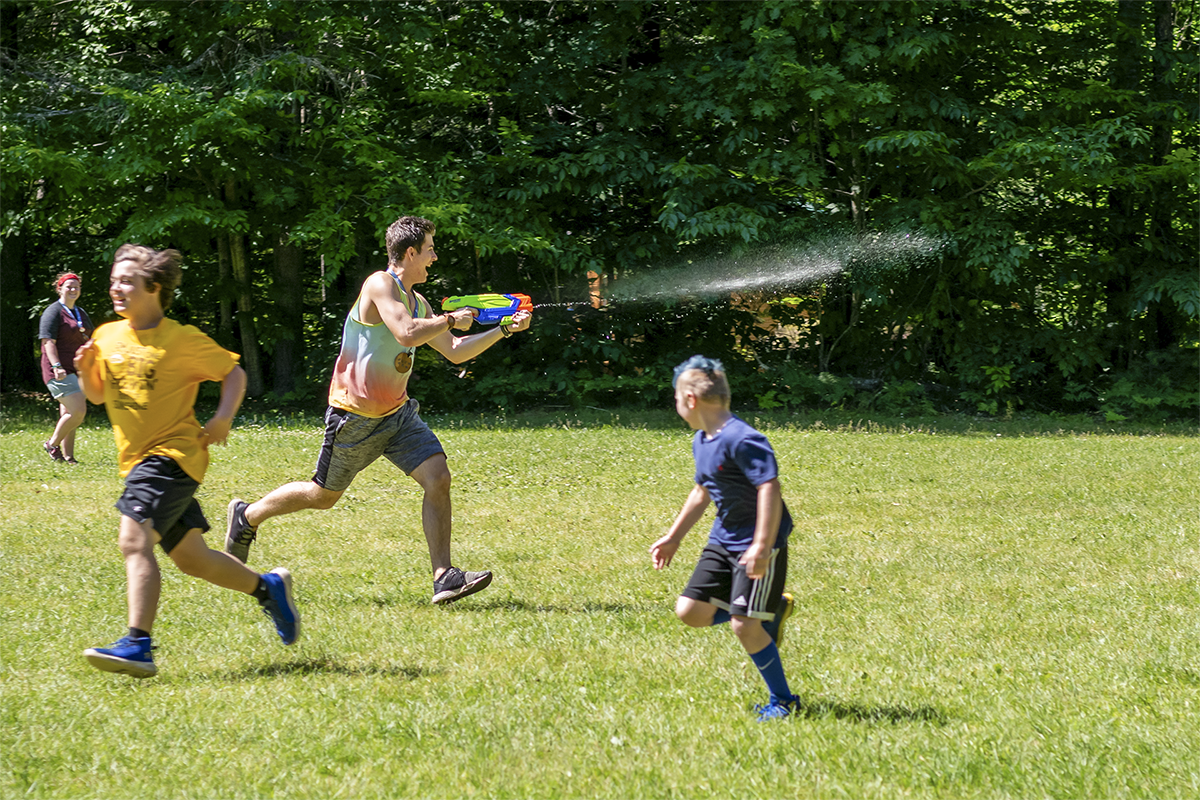 Man in a tie-dye tank top spraying kids with a water gun on a grassy lawn.