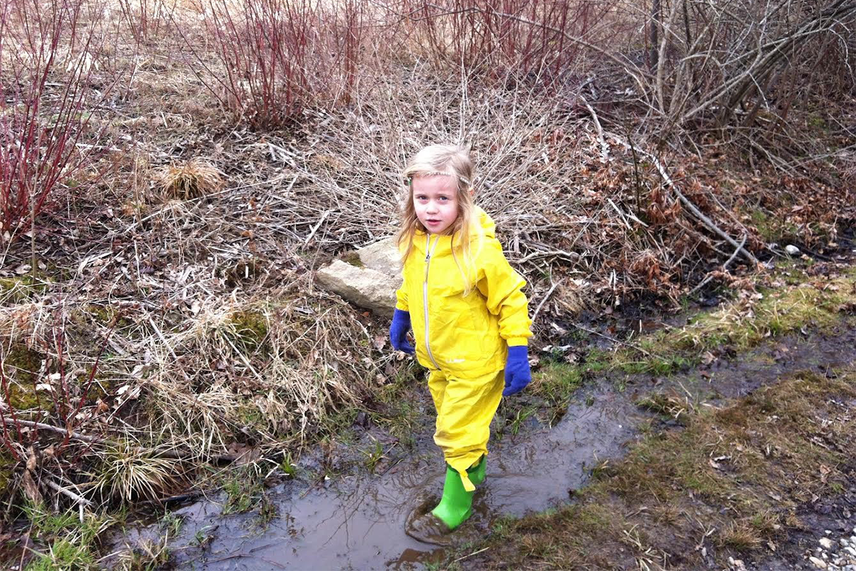 A young blonde girl with green rainboots and blue gloves. She is standing on a muddy trail.
