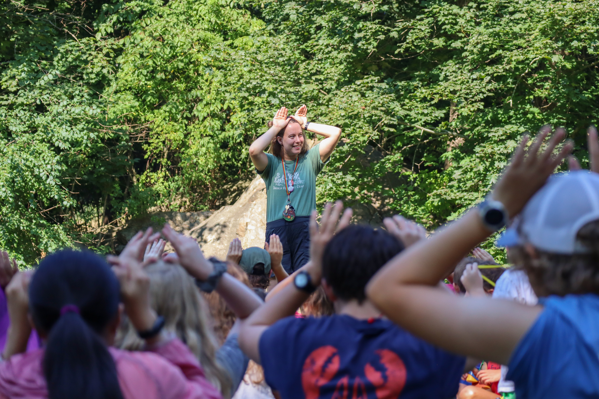 Camp counselor holds their hands at their forehead like bunny ears in front of a group of kids doing the same.