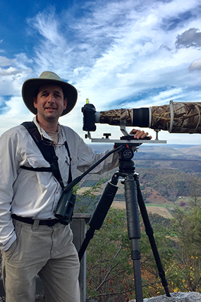 Man with a hat stands smiling next to a large, camouflaged scope.