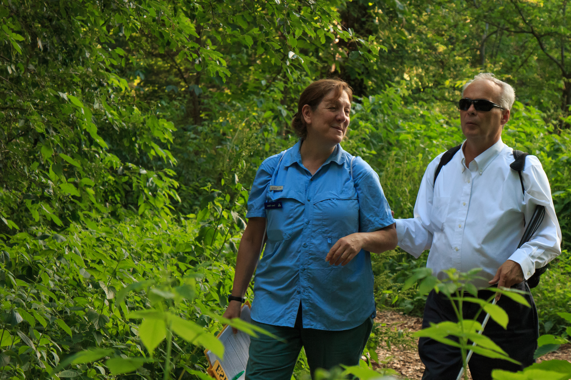 A man (Jerry Berrier) holds the elbow of a woman with one hand and a white cane with the other. They are walking on a path in a green forest.