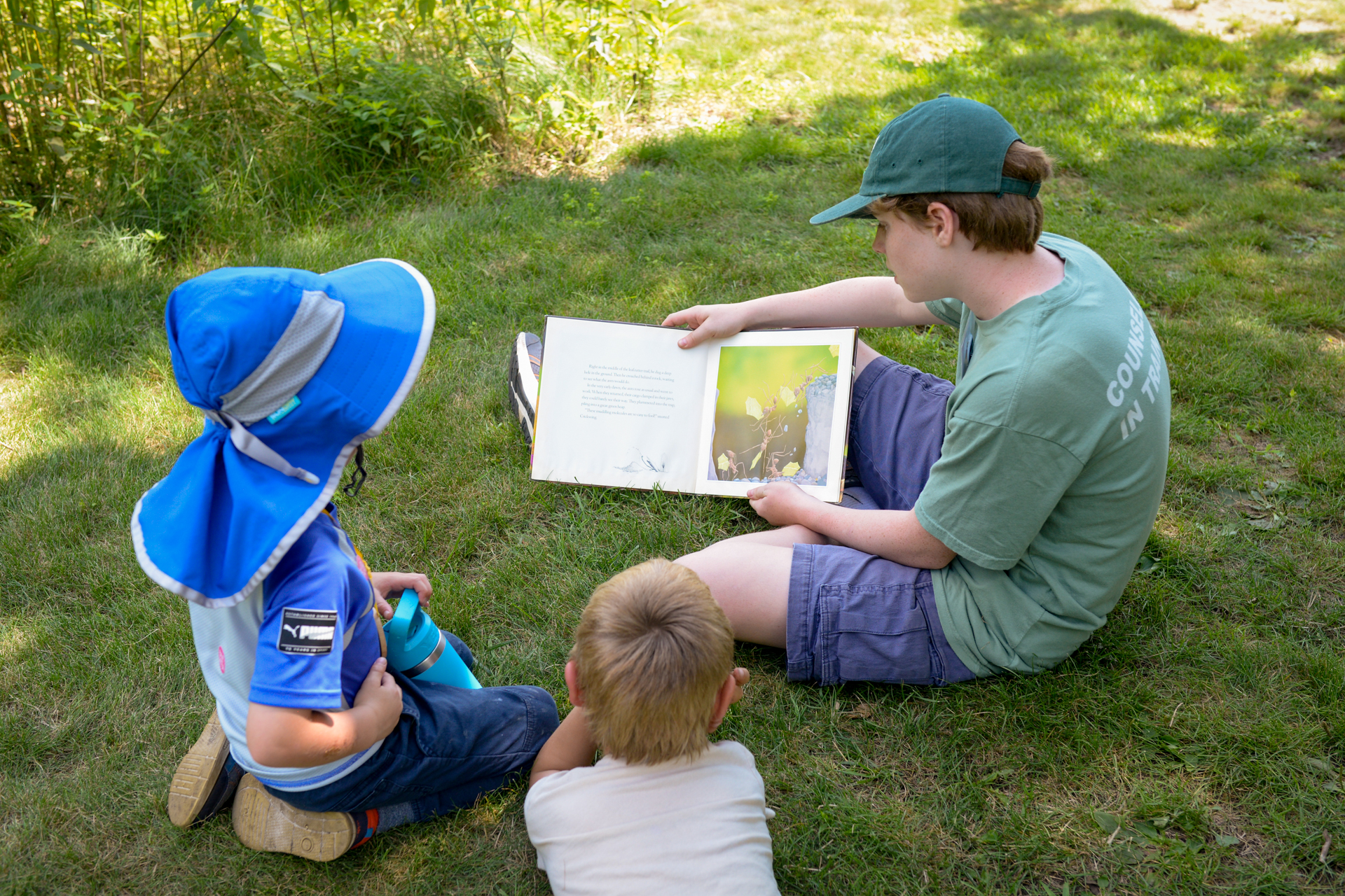 A CIT at Broadmoor Nature Camp reads a picture book to two campers seated on the grass in the shade