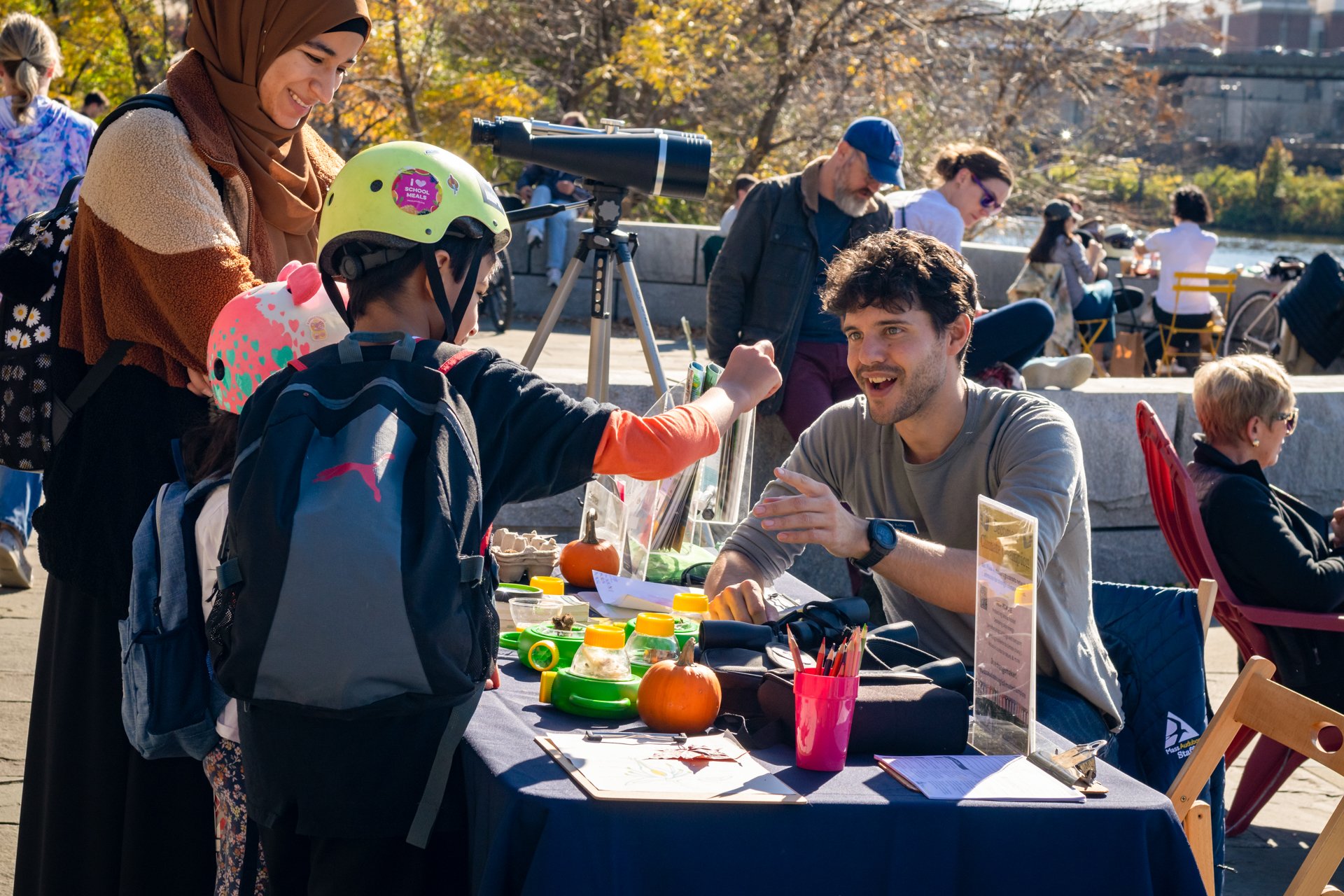 A Mass Audubon educator Magazine Beach engages  with children and a mom at a table covered with kid-friendly microscopes and other nature objects and science tools