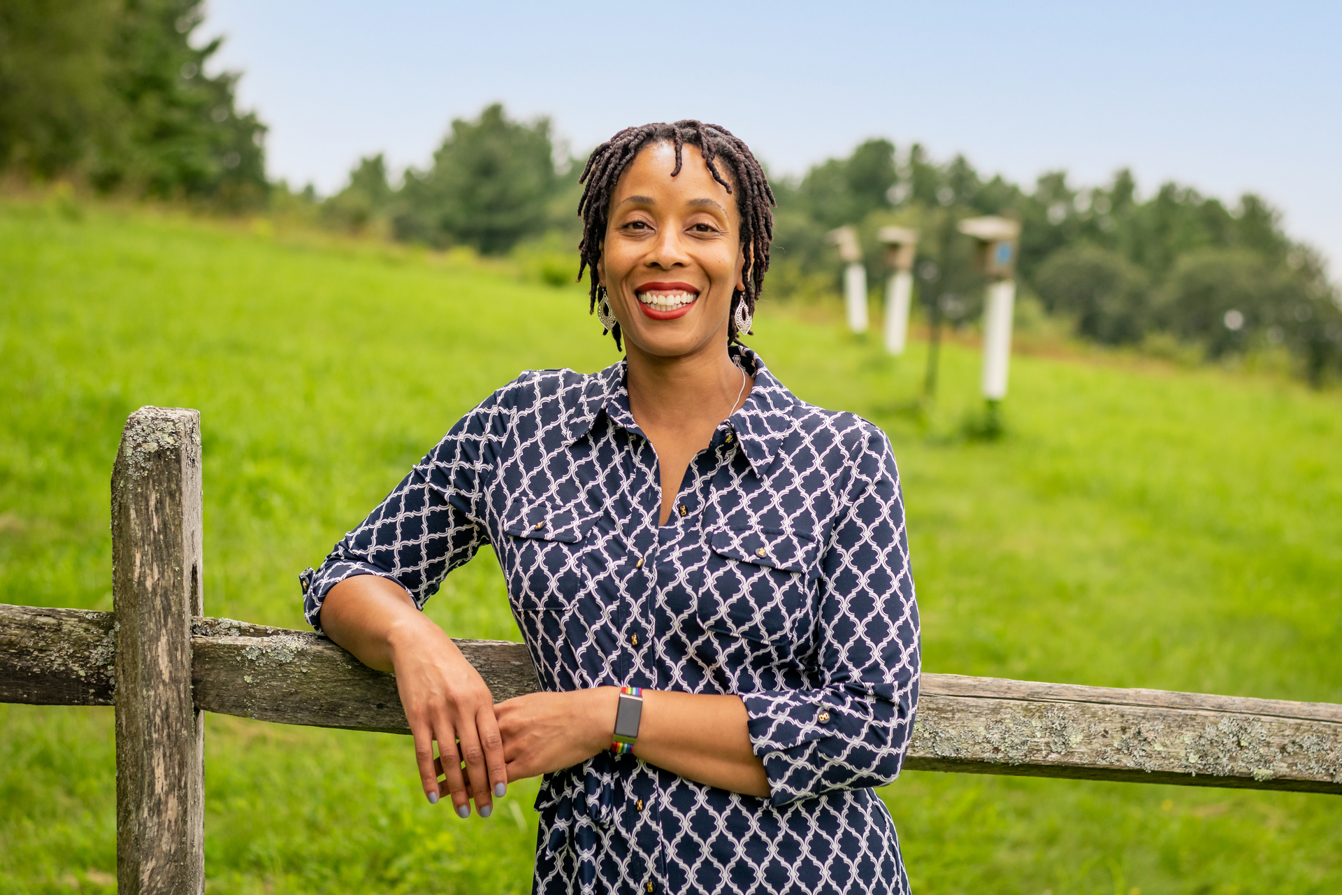 Woman smiling with a blue and white shirt, leaning against a fence with a green pasture in the background.