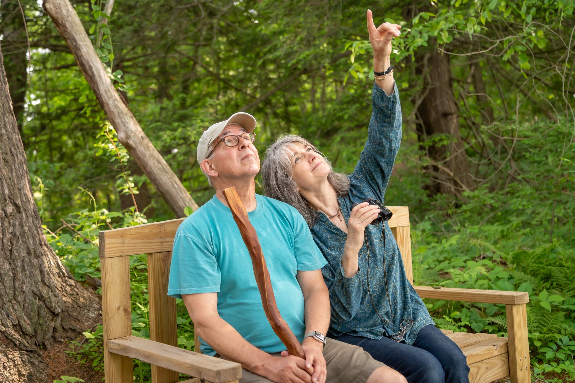 a couple sitting on a bench in the woods, holding binoculars and pointing up.