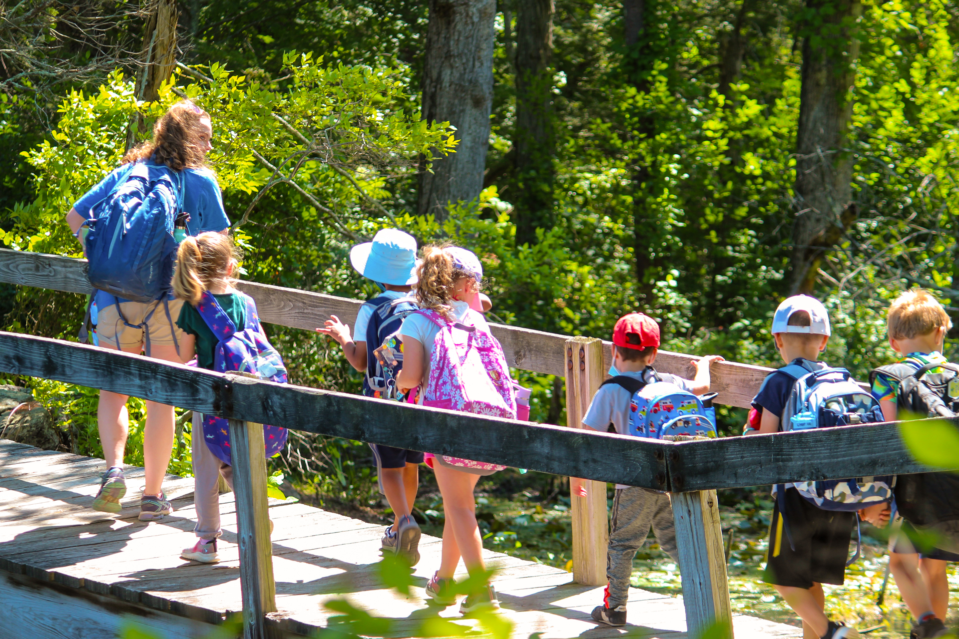 A line of Broadmoor campers wearing colorful backpacks follow their counselor along a wooden boardwalk