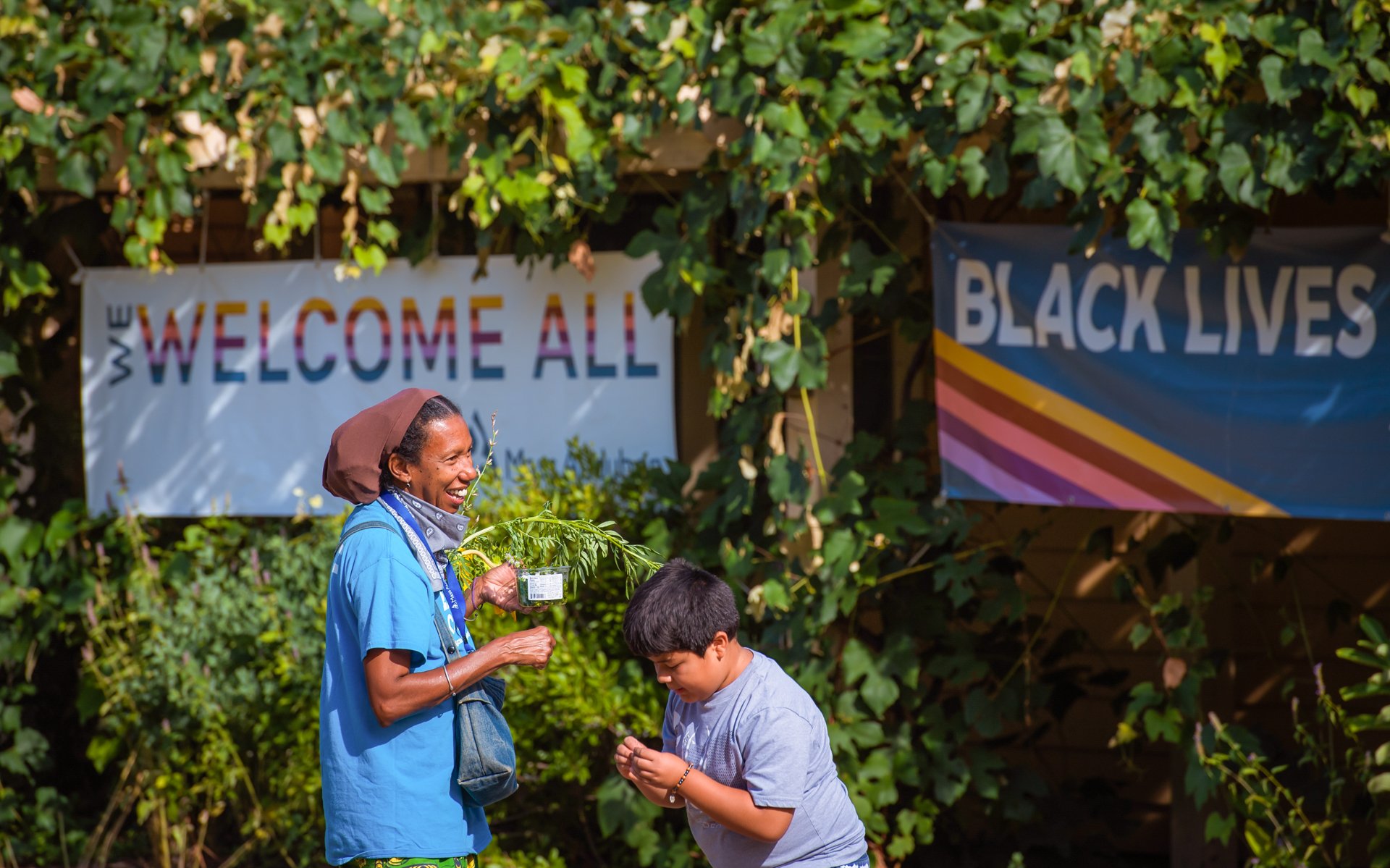 A teacher naturalist and a camper at Boston Nature Center standing in front of two banner signs surrounded by green foliage; one reads "We Welcome All" and the other reads "Black Lives Matter"
