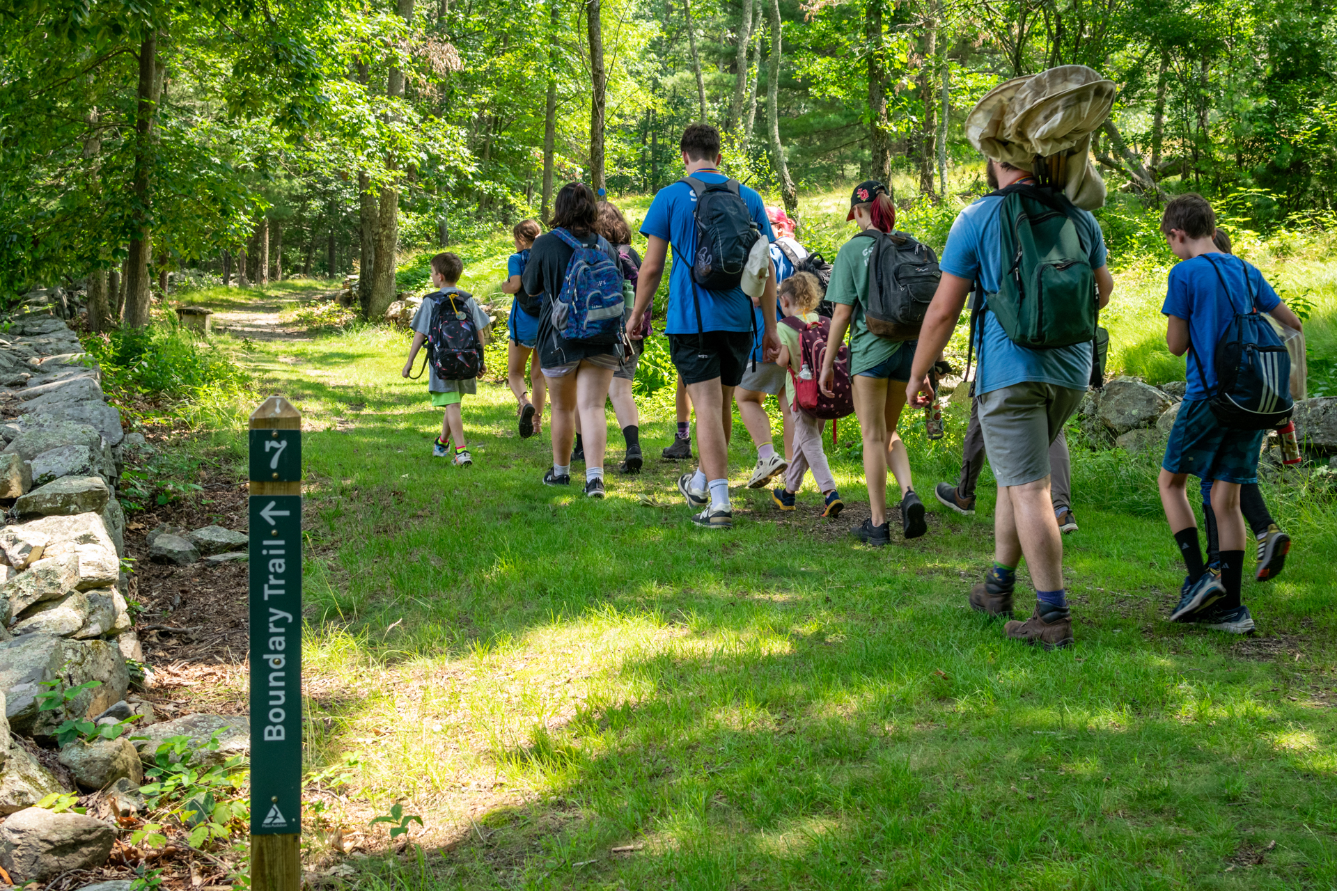 A group of Broadmoor campers and counselors walks along a shady, grassy trail in the woods. A stone wall runs along the left side and there is a trail sign in the foreground that reads, "Boundary Trail"