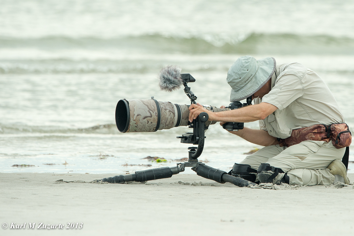 Man knelt down on a sand beach looking into a camouflaged camera scope.
