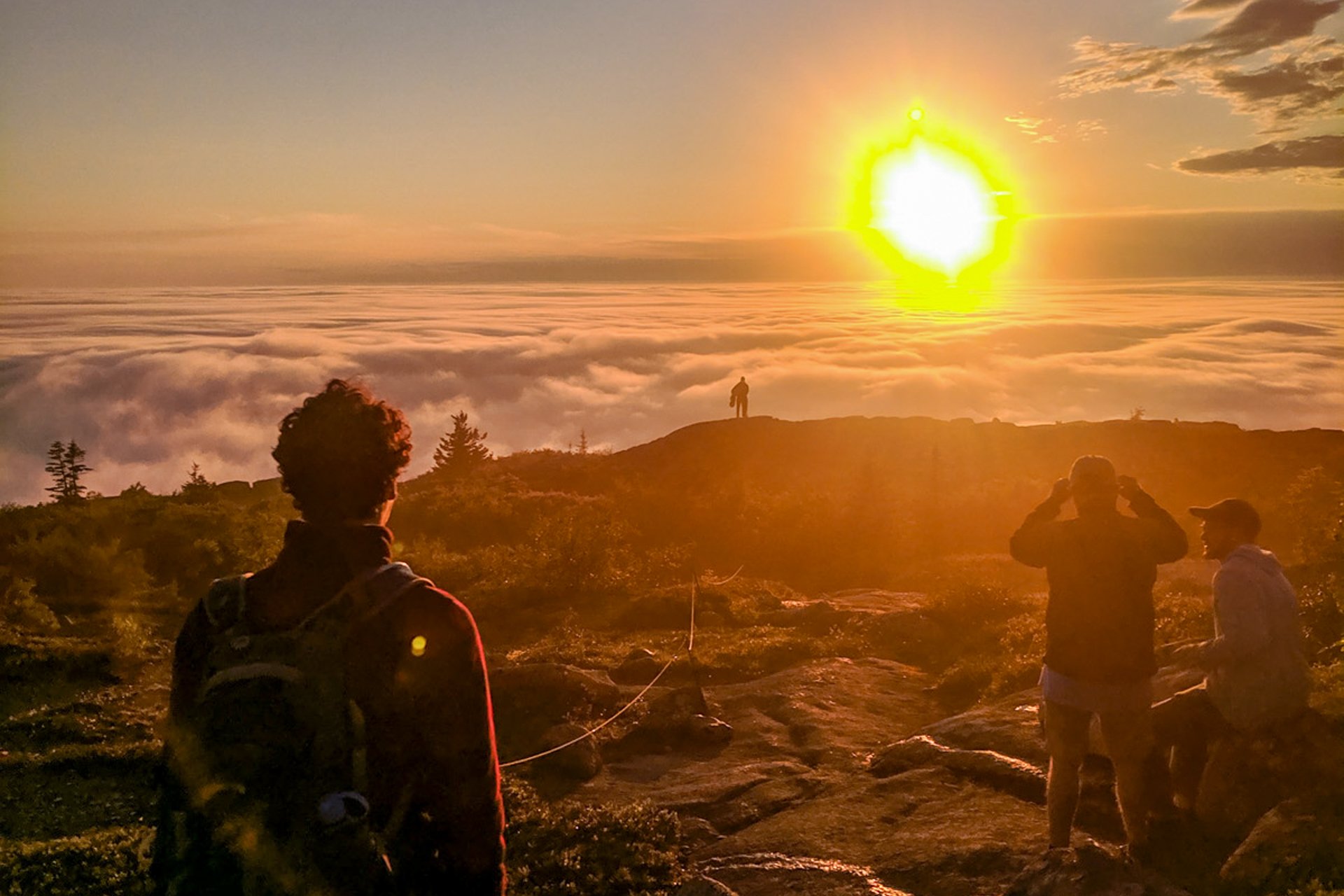 A teen adventure trip participant takes in a stunning sunset from the top of Cadillac Mountain, with a sea of clouds visible below and into the distance