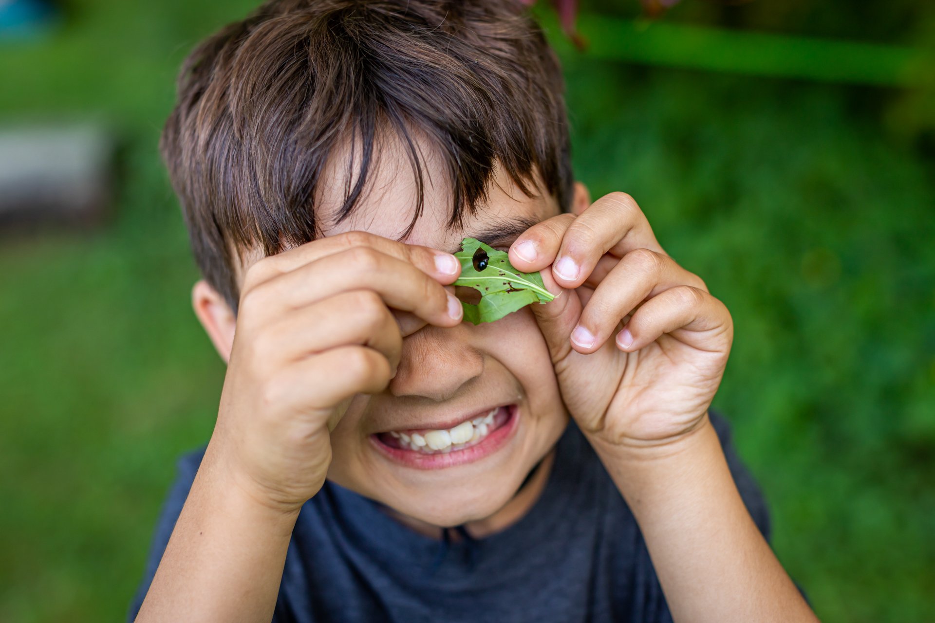 A smiling camper at Arcadia Nature Camp holds a leave up to his eye and peers through the holes made by a hungry insect