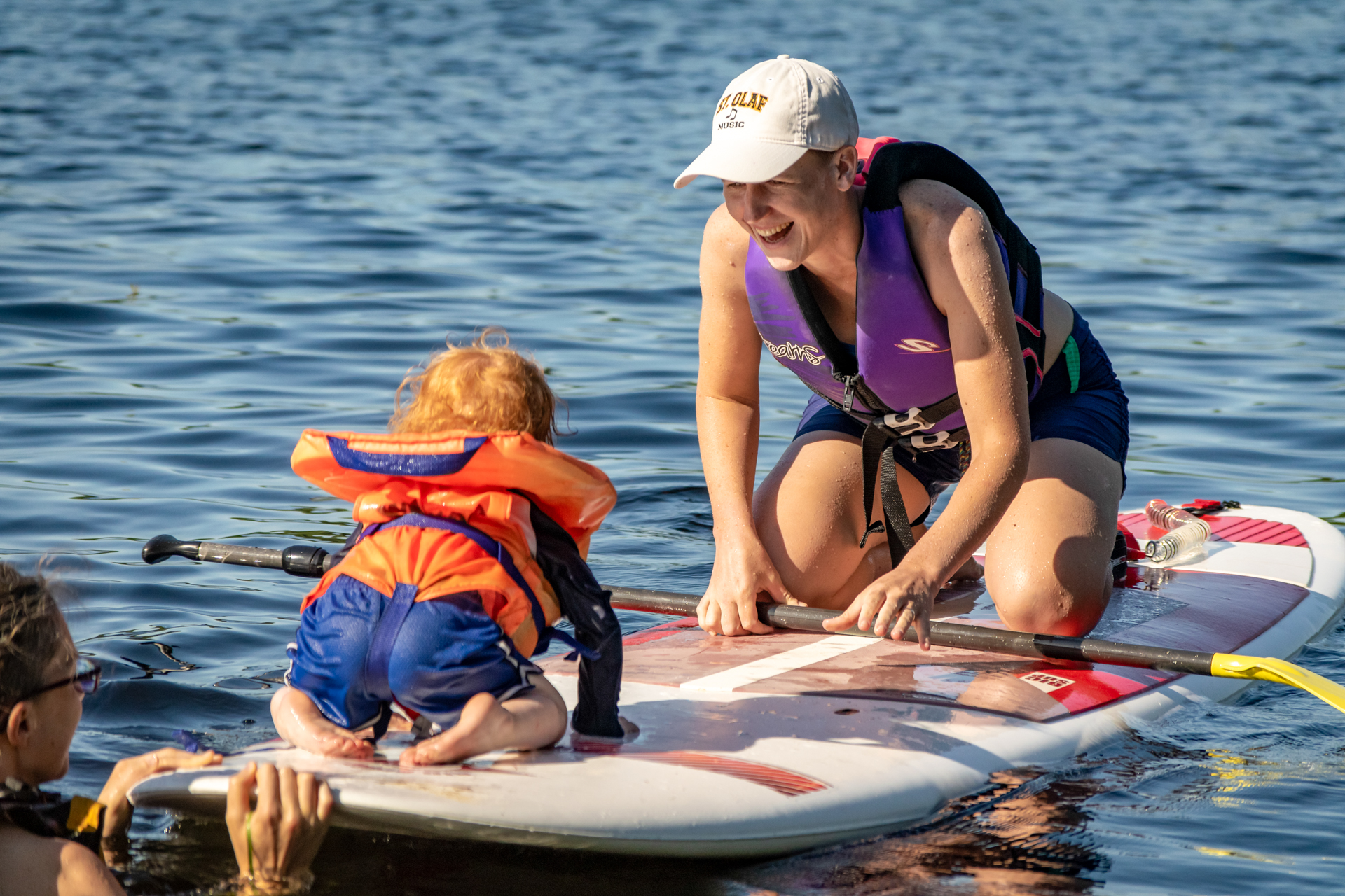 A smiling mom and a toddler, both wearing life jackets, are kneeling on a paddleboard facing each other on calm, blue water