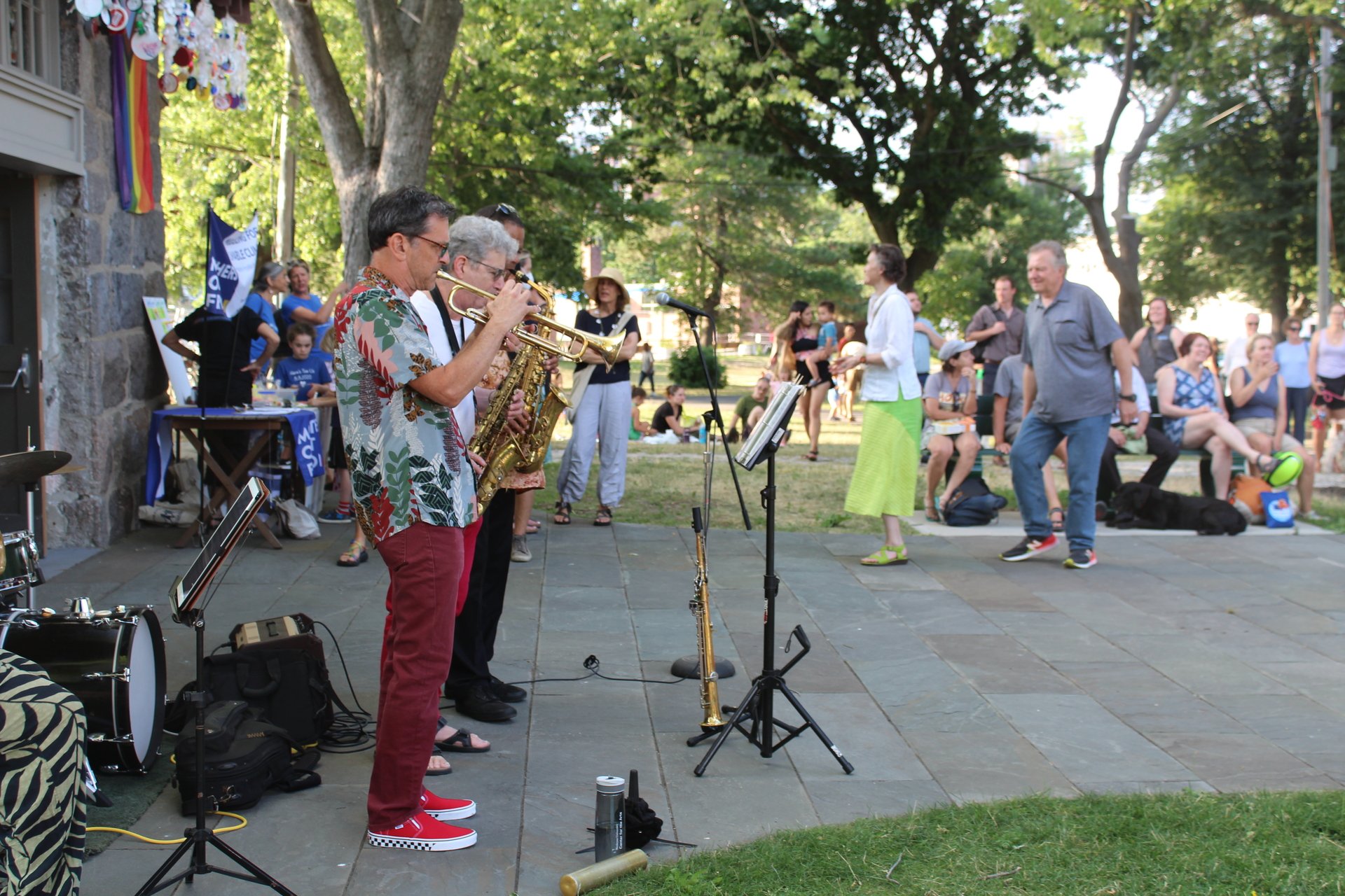 A man in a floral shirt and maroon pants plats the trumpet on a stone walkway. A crowd of people watch and two people are dancing.