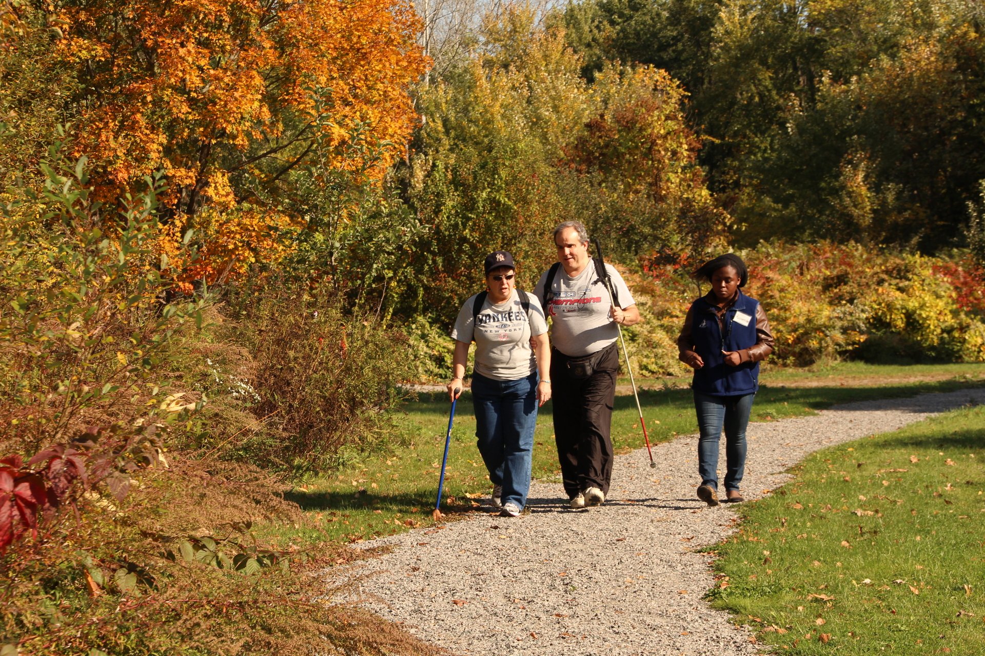 Two people with walking sticks and one without walking on a gravel path towards a the camera.