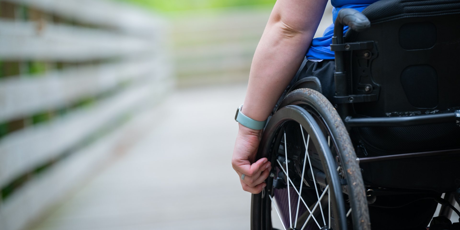 A close-up, cropped photo of a person seated in a wheelchair with their hand on the wheel, using the accessible All Persons Trail boardwalk