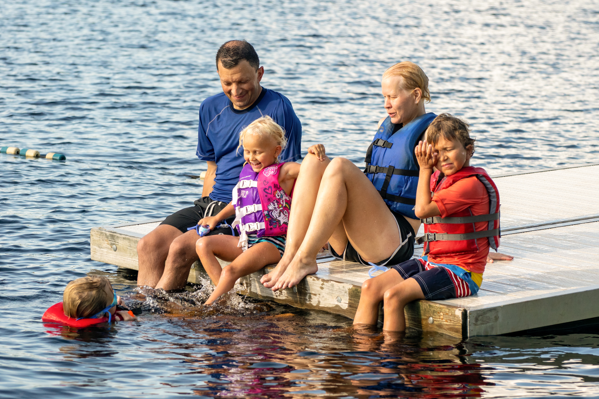 At Wildwood's Family Camp, a mom and dad and their three kids are seated on the edge of a dock, dangling their feed in the water. One child is in the water swimming; all three are wearing life jackets.