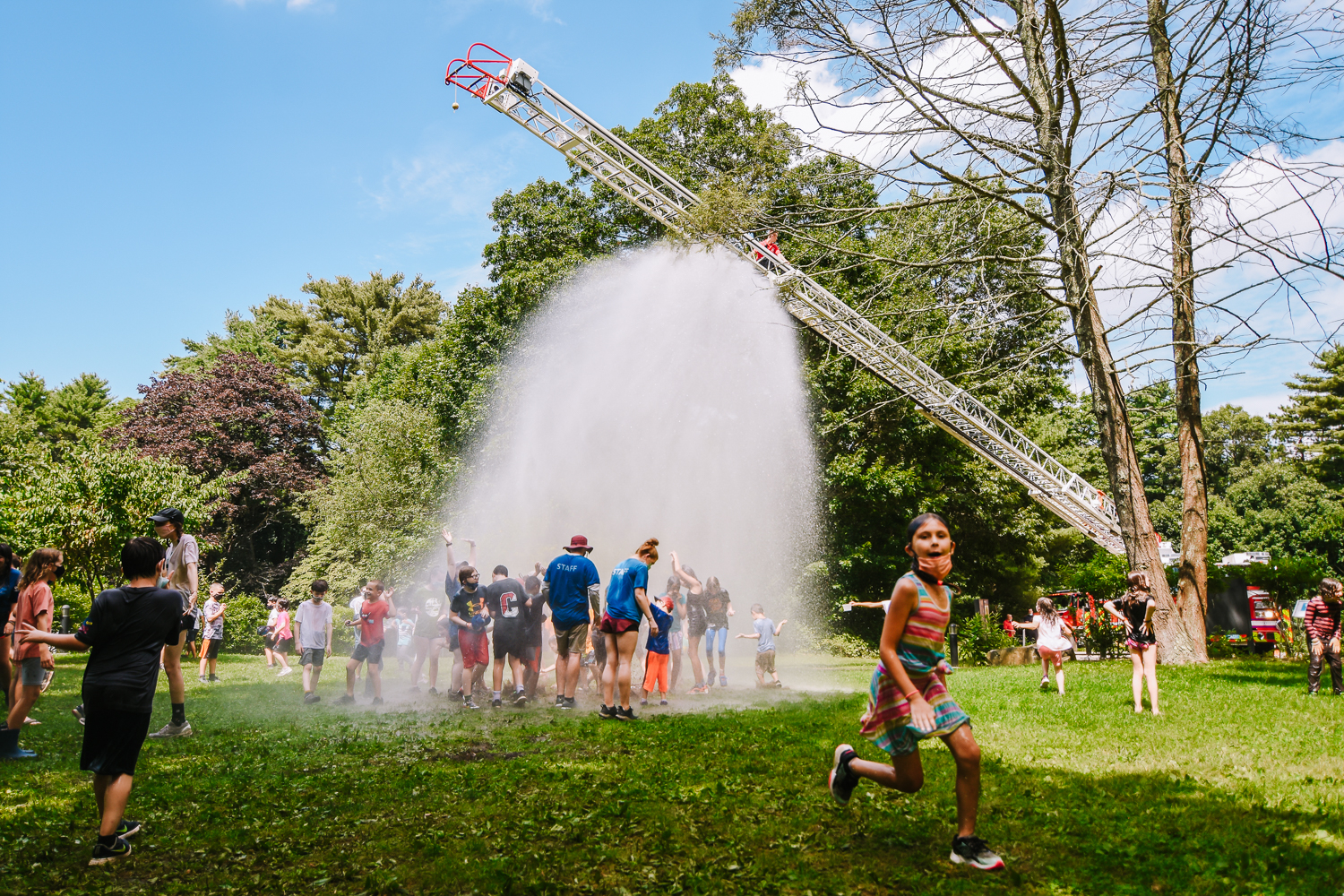 A firetruck with its ladder extended sprays a shower of water from its hose over a field full of campers on a hot day at Moose Hill Nature Camp