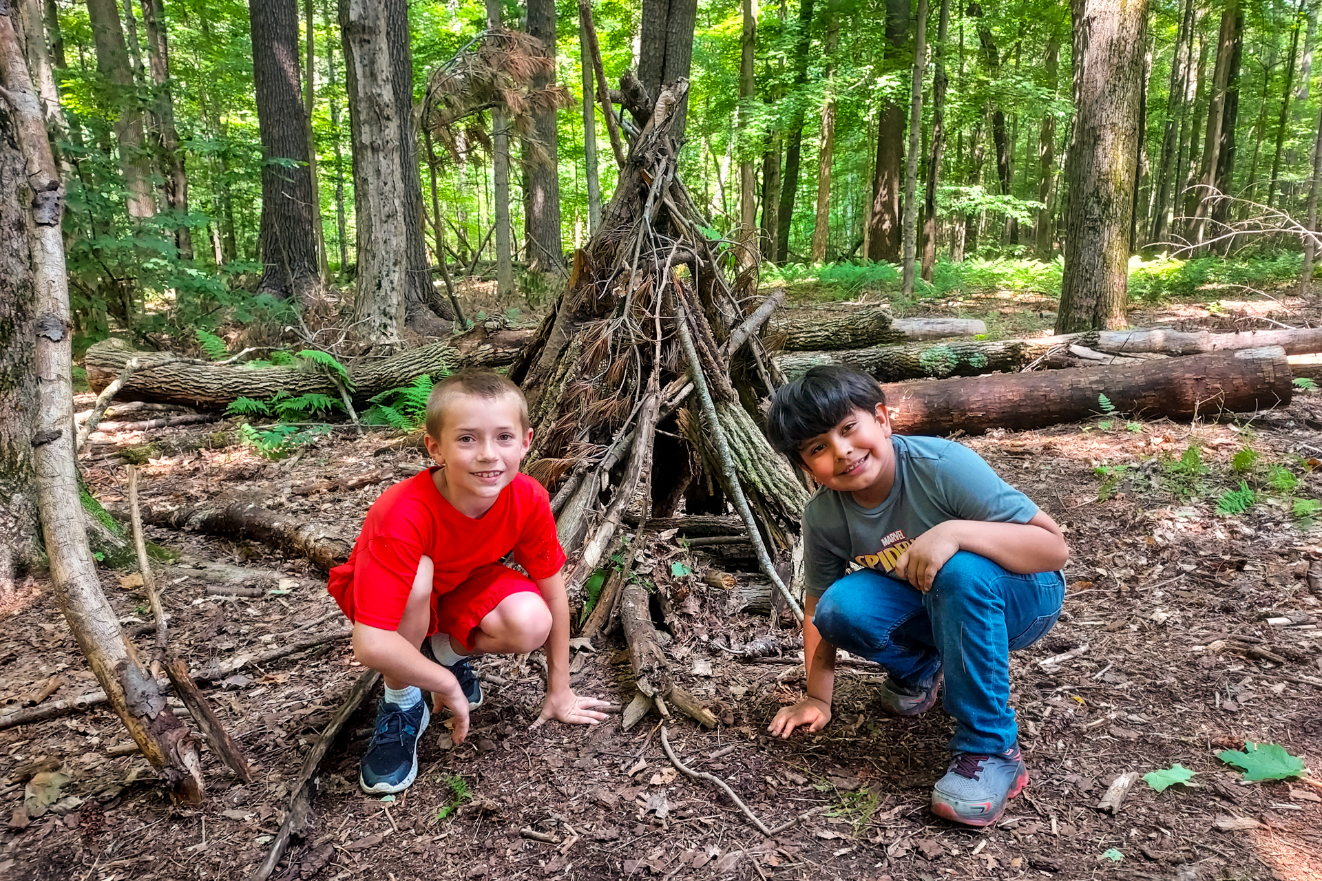 Two campers at Berkshire Nature Camp at Pleasant Valley kneel in front of a shelter they've built from branches and other natural objects