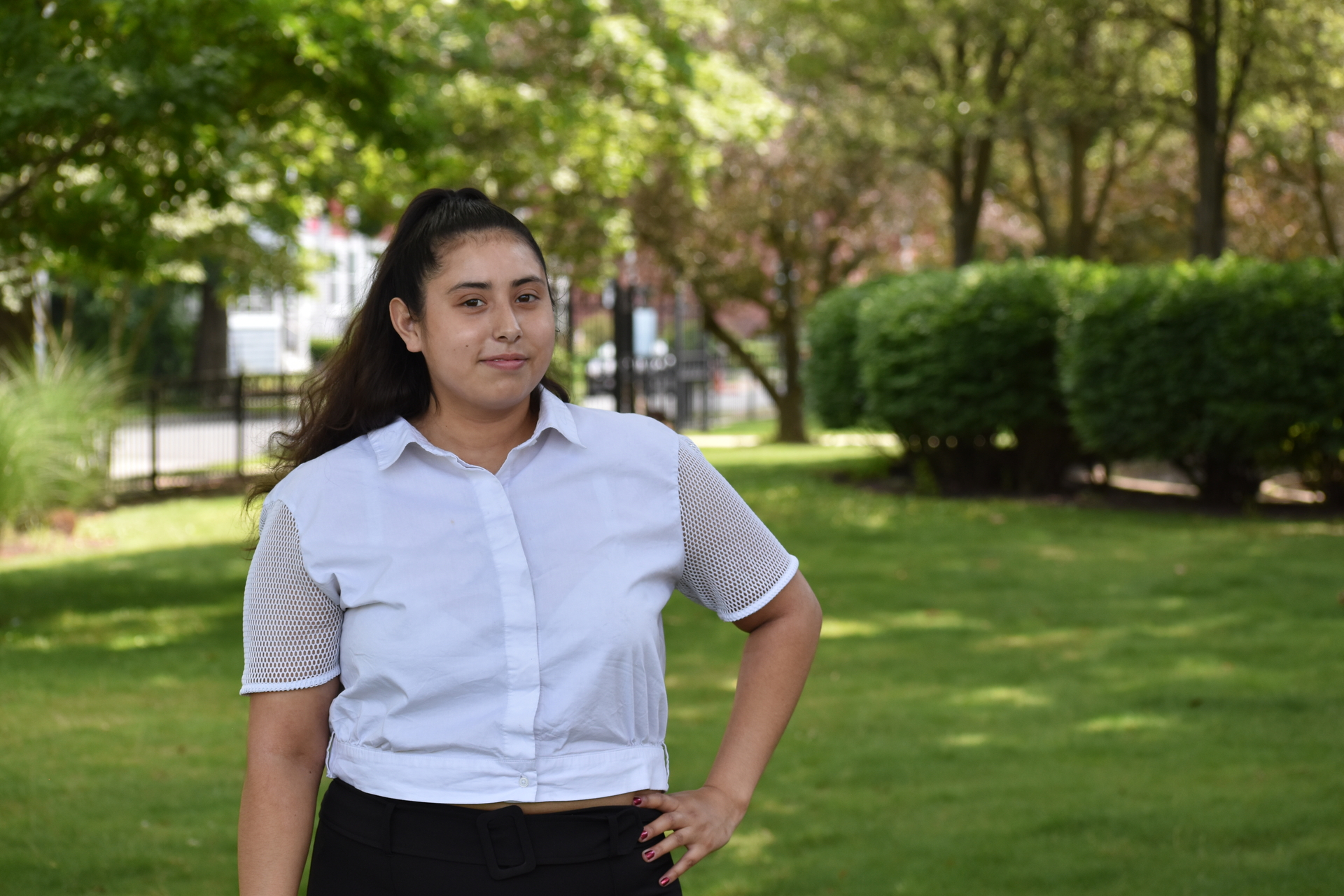 A girl stands in a park with a white shirt and one hand on her hip.