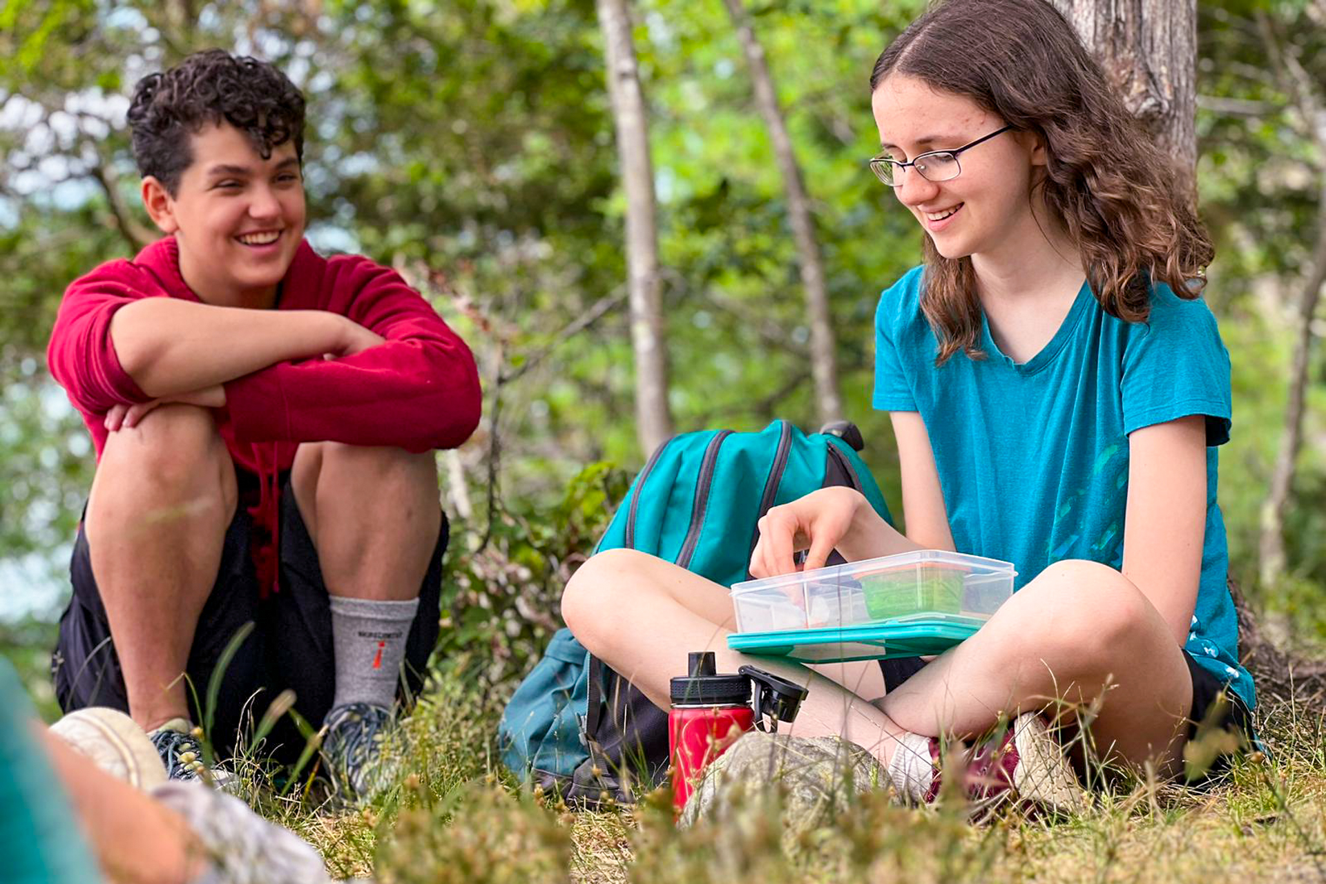 Two teenage campers at Moose Hill Nature Camp sitting in the grass, eating lunch and laughing