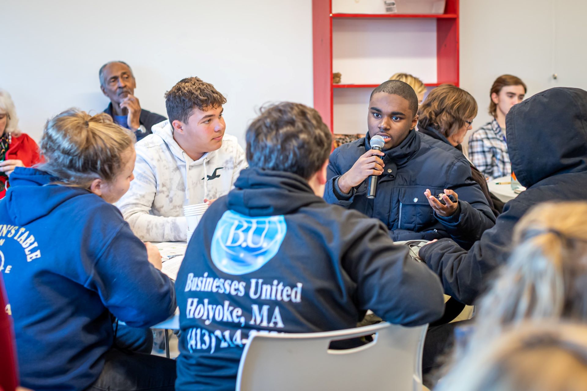 student seated at table speaking into microphone in a group of high school-aged students.