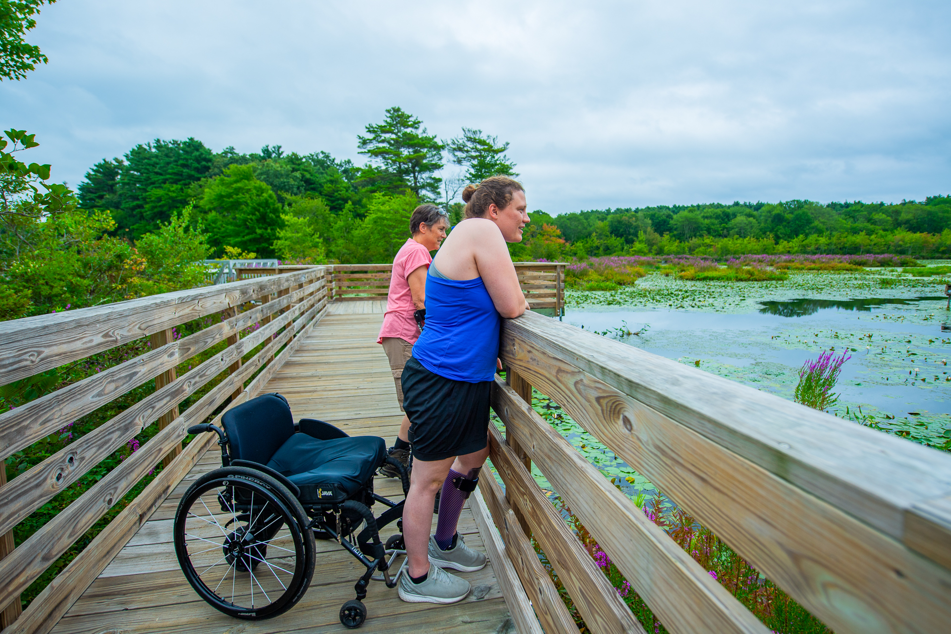 Two people leaning against the railing on Stony Brook's wheelchair-accessible All Persons Trail; one is using arm crutches and one has a wheelchair but is standing to see over the railing