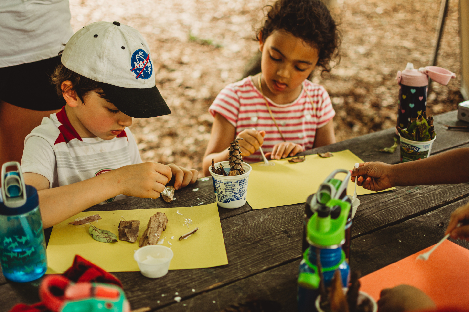 Two campers at Moose Hill Nature Camp making art with construction paper, glue, and natural objects like bark, leaves, and cones