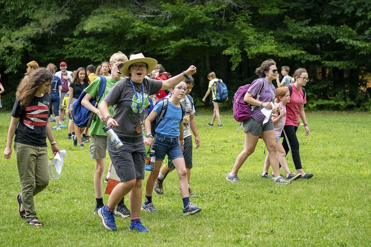 Person with a tan hat walks in front of a group of children with one hand in the air.