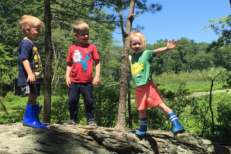 Three campers in mud boots climbing a rock in the sunshine at Broadmoor Nature Camp